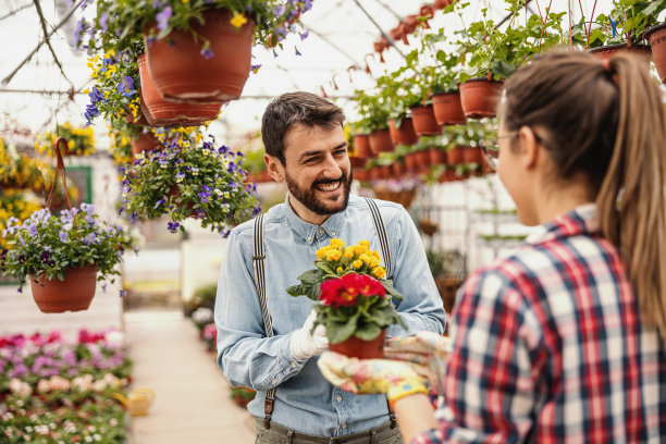 两个微笑着相爱的企业家，一边调情，一边捧着花盆和鲜花。周围都是五颜六色的花朵。温室内部。图片下载