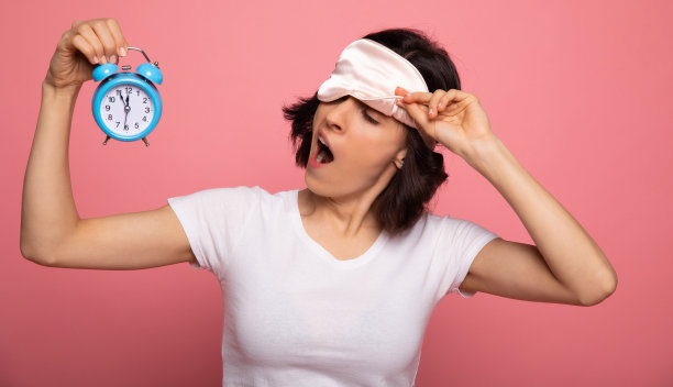 沉重的卧铺。一个疲倦的女人穿着口罩和睡衣的特写照片，谁是打哈欠，而看在她的右手闹钟。图片下载