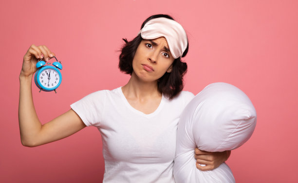 不满意。这是一张漂亮女孩的特写照片，她头上戴着睡眠面具，手里拿着一个闹钟和一个枕头，绝望地看着镜头。图片下载