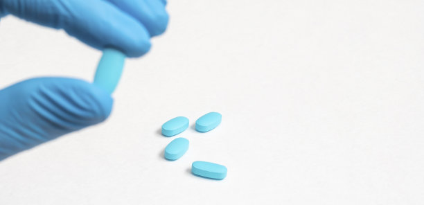男性医生的手在蓝色消毒手术手套拿着蓝色胶囊在白色背景与几个蓝色药丸在背景的特写。模糊前景，聚焦背景。健康理念旗帜图片下载