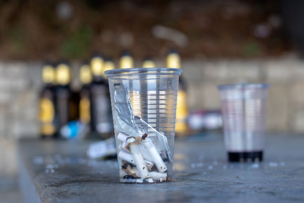 晚上在公共场合喝酒后留在公园里用作烟灰缸的塑料杯。酒瓶在背景中图片下载