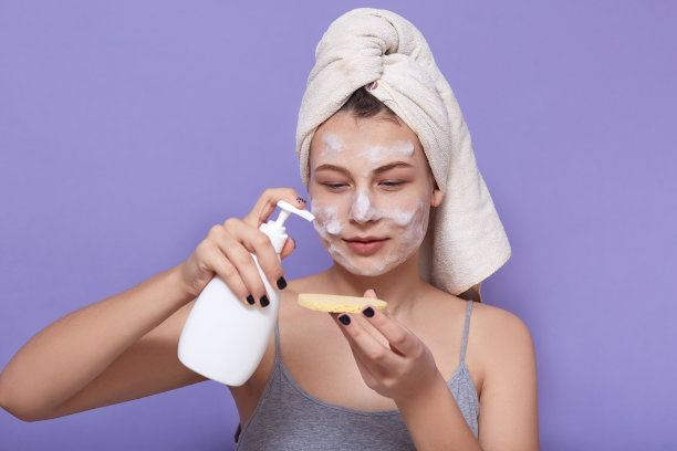 这是一幅室内照片，年轻女性在海绵上涂护肤霜，在淡紫色的背景下摆出孤立的姿势，女性专注于海绵上的凝胶或乳液，在家里做晨妆。图片下载