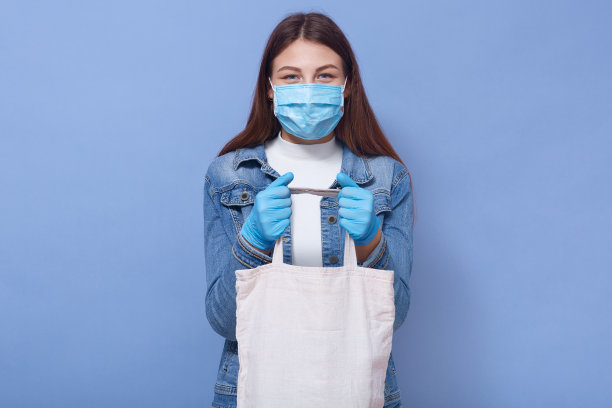 水平摄影棚照片，细心的年轻女性直视着镜头，拿着装有物品的环保袋，戴着防护口罩和手套，在检疫期间购物。Covid 19的概念。图片下载