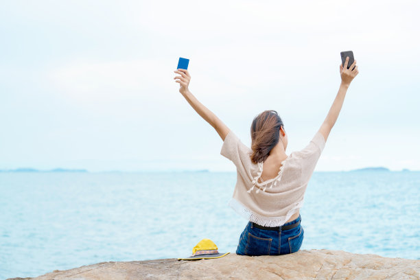 年轻美丽的亚洲女人女孩的后视图坐在岩石海岸的海滩上拿着智能手机和信用卡，在空中伸展她的手臂与网上银行成功购物图片下载