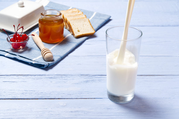 往玻璃杯里倒牛奶。一杯牛奶，蜂蜜和面包，浅蓝色的木头背景。乡村早餐食品概念。图片下载