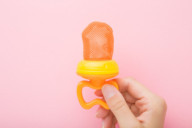 年轻的母亲手里拿着橙色的咬饵，放在淡粉色的桌子上。婴儿咀嚼新鲜水果或浆果的附件，通过网袋喂食器。柔和的颜色。特写镜头。自顶向下的观点。图片下载