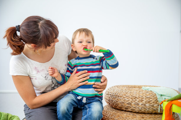 小男孩学习如何正确刷牙，而坐在他的母亲的腿上，对白色的背景-库存照片图片下载