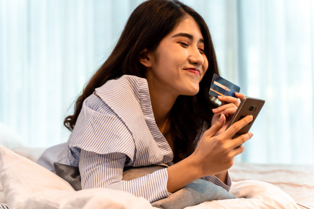 一个快乐的20多岁的亚洲妇女在家中的床上穿着休闲衣服拿着信用卡和手机的肖像。金融、电子商务、网上理财概念图片下载