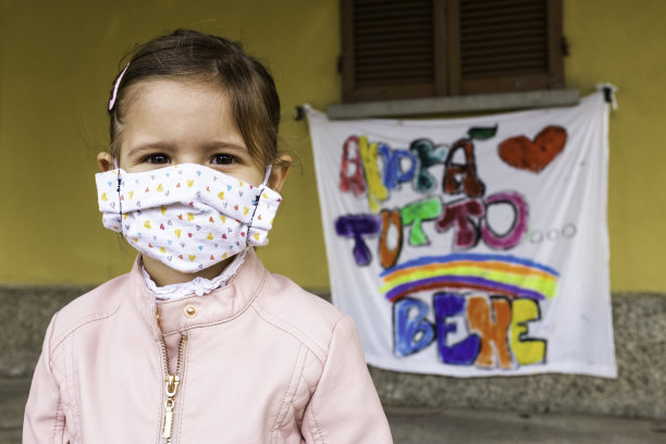 小女孩戴着自制的防护面具。模糊的背景与著名的彩虹画和意大利语的信息“一切都会好起来”。重新启动社会生活。的童年。希望。图片下载