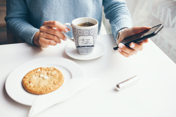 女人手握手机的特写，而喝杯茶和吃燕麦饼干在现代咖啡馆。图片下载