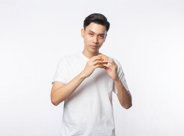 年轻的亚洲男子在白色t恤拿着汉堡，看着镜头和微笑孤立的白色背景图片下载