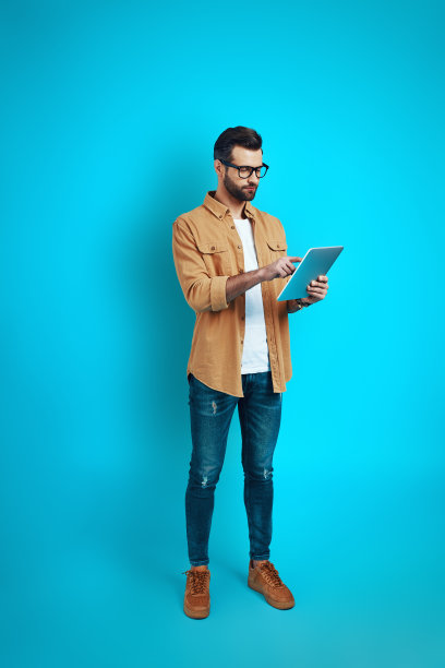 全长度的年轻人在智能休闲装使用数字平板电脑，而站在蓝色背景图片下载