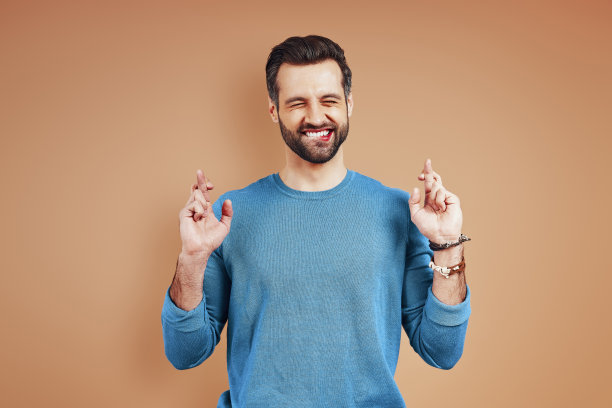 快乐的年轻人穿着休闲装保持手指交叉和微笑，而站在棕色的背景图片下载