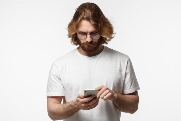 室内照片，一个戴着圆眼镜的大胡子年轻人，在灰色背景下看着智能手机，给他的朋友发信息图片下载