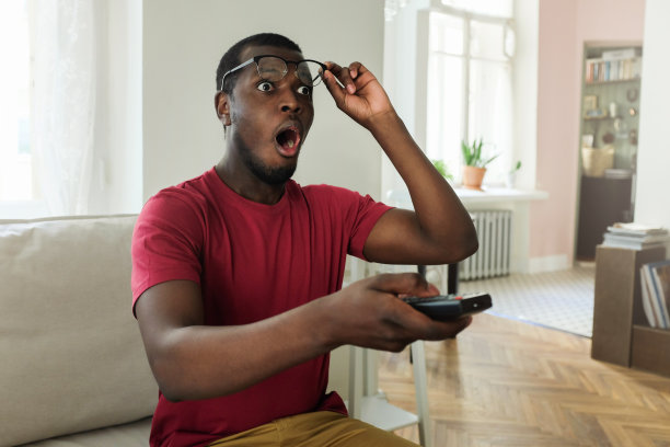 水平图片的年轻非洲男子花空闲时间在家里看电视，看着惊讶和震惊与张开的嘴举起眼镜，以确保他看到的屏幕是真实的图片下载