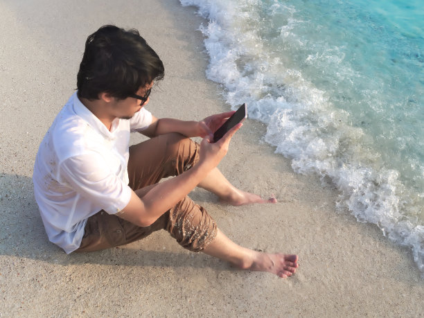 放松的年轻亚洲男子与移动智能手机坐在热带海滩的沙子。暑假的概念。图片下载
