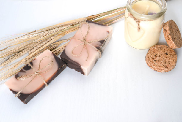 由两个手工制作的带有橙色色调的棕粉色肥皂，柔和的香草蜡烛和小麦穗组成的白色背景和木质纹理。水平的照片。手工化妆品概念与天然成分的个人护理，洗澡图片下载