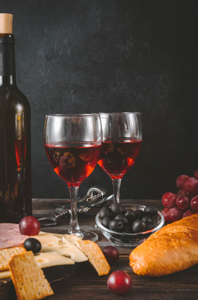 2杯红酒配开胃品，瓶子，奶酪片，火腿，果酱，葡萄，橄榄，法式长棍，饼干，深色木头上的开瓶器图片下载