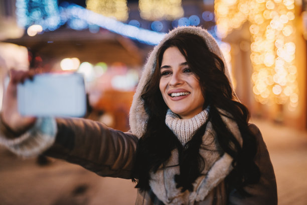 美丽微笑的女孩在冬天的大衣站在街上和她自己的照片。城市街道上的圣诞装饰。冬天的夜晚。图片下载