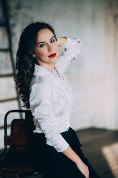 一个漂亮的棕色眼睛的小女孩，一头卷发，红唇，穿着白衬衫和黑裤子，坐在录音室的酒吧凳子上。软选择焦点。图片下载