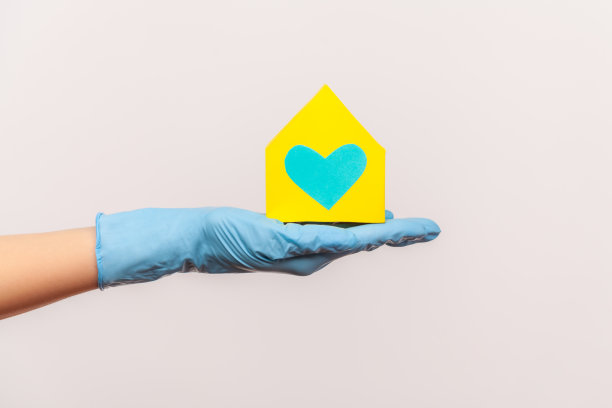 侧面特写人类的手在蓝色的外科手套拿着黄色的纸房子外观在手与爱和照顾。图片下载
