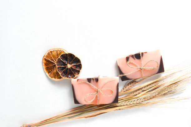 平铺的构图是两种带有橙色色调的手工制作的棕粉色肥皂，在白色背景上有小麦穗状花序的干橘子片，带有木质纹理。位于画面右侧的物体。水平照片，复制空间图片下载