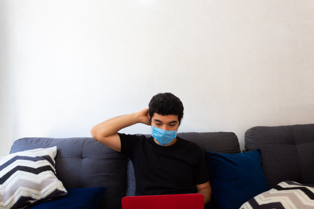 一名戴着蓝色医用口罩的年轻人因新冠肺炎隔离在家使用笔记本电脑工作图片下载