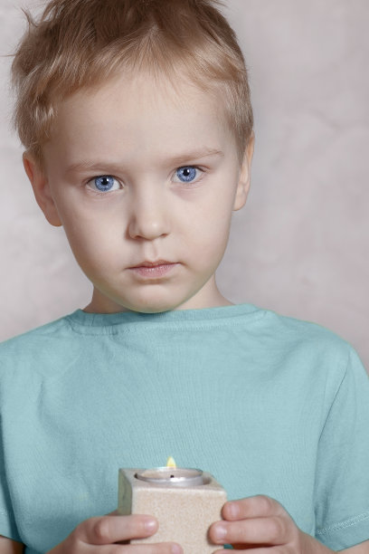 美丽而严肃的白人小男孩看着镜头，双手捧着燃烧的蜡烛。图片下载