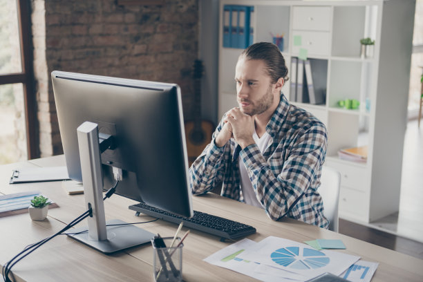 英俊的业务经理小伙子的照片，看电脑显示器，桌子，认真读公司报告，穿休闲衬衫，西装，坐椅子，现代的室内，宽敞的办公室，室内图片下载