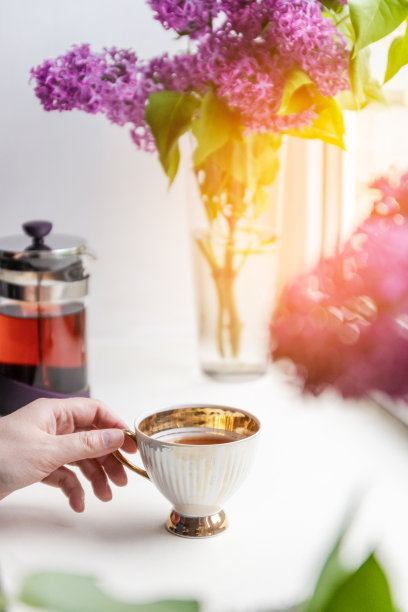 在一个阳光明媚的日子里，窗台上的一个玻璃花瓶里放着一个瓷杯，里面盛着热茶和一束丁香花。垂直图图片下载