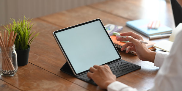 裁剪图像的图形设计师的手拿着一支手写笔和打字在电脑平板电脑与键盘的情况下，而坐在一个有序的工作场所的木制办公桌作为背景。图片下载