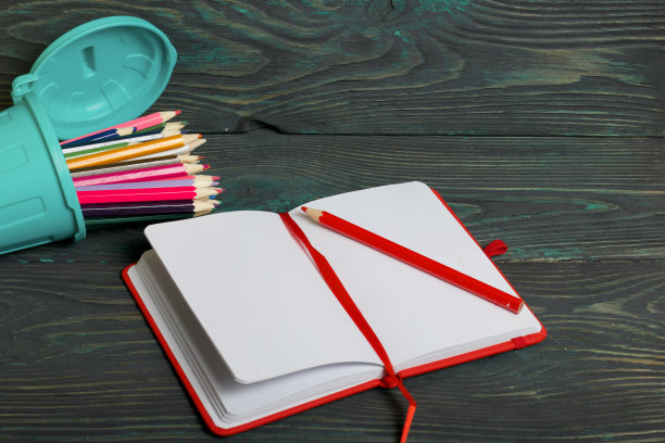 一只装着彩色铅笔的垃圾桶侧面放着一支铅笔。靠近一个打开的笔记本。背景是刷成黑色和绿色的松木板。图片下载