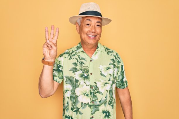 一位灰白头发的中年男子戴着夏帽，穿着花衬衫在海滩度假，一边自信而快乐地微笑着，一边伸出第三个手指。图片下载