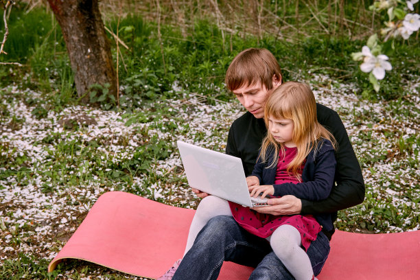 一个女孩和一个男孩背靠背坐在笔记本电脑后面，在一个花圃里学习。以绿草和开花的树木为背景。远程娱乐。图片下载
