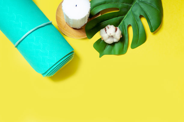 天然木制彩色蜡蜡烛与棉花，绿色的叶子和卷绿松石薄荷瑜伽，普拉提垫在黄色的背景。顶部视图和文本空间。放松和冥想的概念。图片下载