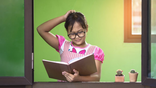 戴眼镜的亚洲黑皮肤小女孩在家里的玻璃窗框里阅读课本时，用怀疑和怀疑的手势挠着头图片下载