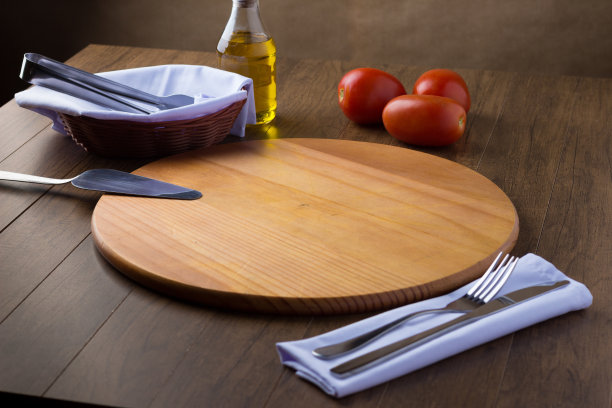 圆木板，三个红番茄，篮子和餐巾和支架，钢抹刀，杯子和橄榄油，刀叉餐巾。在美食区用来供应披萨的餐具。曝光不足的照片。图片下载