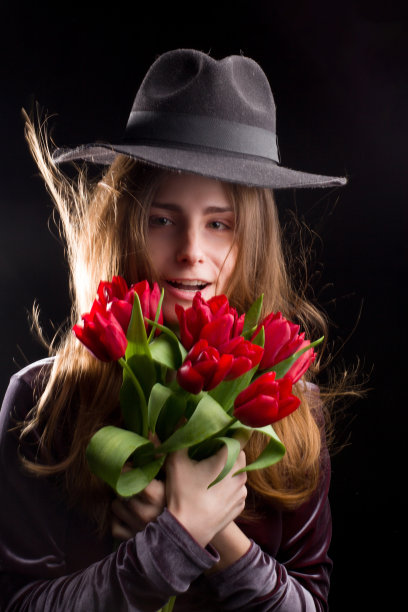 画室里，深色背景上，身着紫色礼服，头戴黑色宽边帽的女孩，手里拿着一朵红色郁金香花，闭上了眼睛图片下载