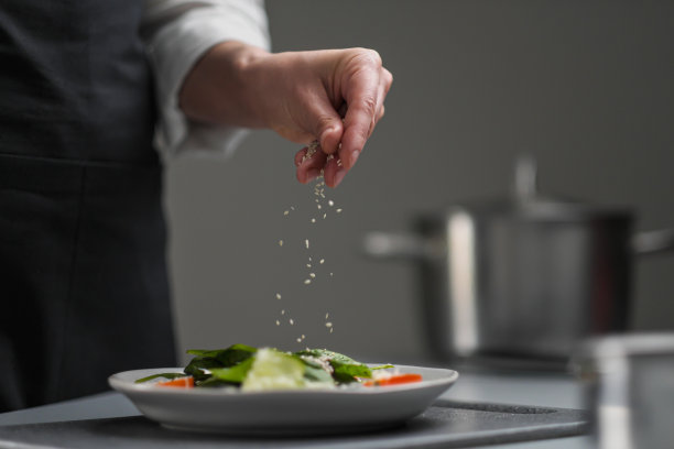 一个穿着白色制服和黑色围裙的女厨师在餐厅厨房里。厨师在沙拉上撒上芝麻。图片下载