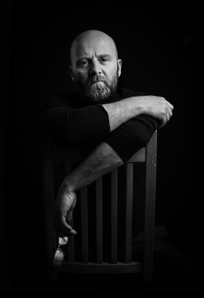 一个留着胡子的秃顶男人的简单画室肖像，胳膊随意地搭在椅背上，一只手里拿着一副眼镜，捕捉着光线。黑色背景，便于扩展复制。图片下载