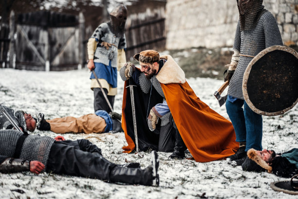 一个穿着锁子甲和披风的中世纪战士跪在战死的士兵面前。以冬天的森林和雪为背景图片下载