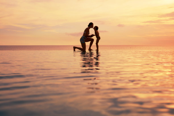 家庭。母亲和孩子的剪影。妈妈和女儿在海滩上牵手。美丽的落日天空和水中倒影。图片下载