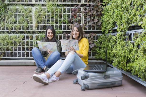 两个漂亮的女人在看地图。绿色背景。他们笑着，寻找一个度假的地方。户外生活方式和友谊。还有手提箱和笔记本电脑。图片下载