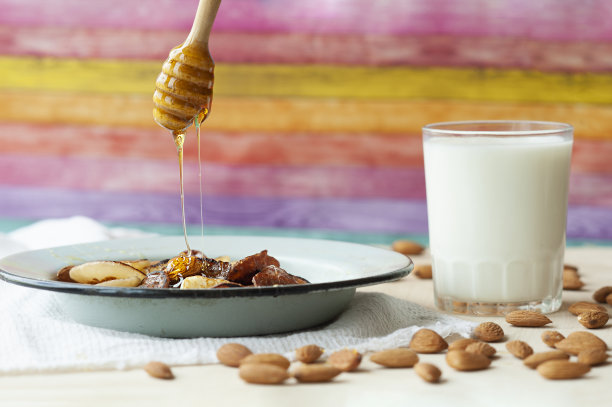 蜂蜜从一根木棍上滴在小煎饼上，上面有杏仁和蜂蜜，放在色彩明亮的厨房里，还有牛奶杯和散落的坚果。trandy食物照片图片下载