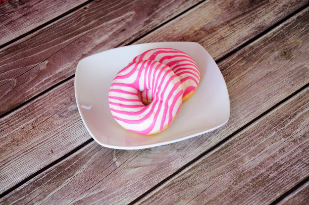木桌上放着白色陶瓷盘子，上面放着两个新鲜的粉色圆形甜甜圈。图片下载
