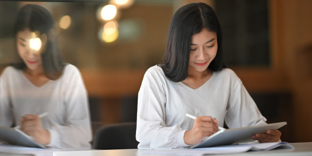 美丽的女人拿着一个电脑平板电脑和手写笔的照片，而坐在那里，看着它在舒适的客厅作为背景的白色办公桌。图片下载