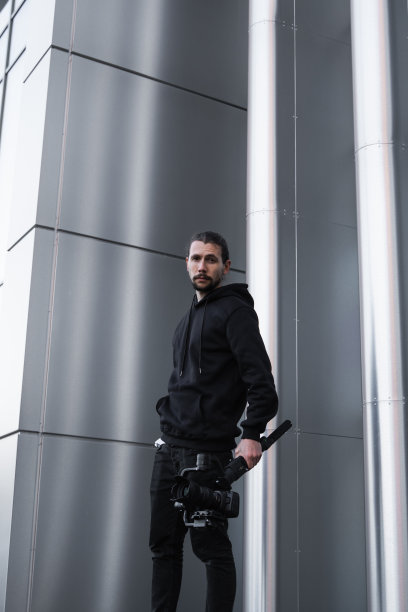 年轻的专业摄像师手持专业摄像机，安装在三轴框架稳定器上。专业设备有助于制作高质量的视频而不摇晃。一个穿着白色连帽衫的摄影师正在录像。图片下载