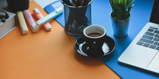 咖啡杯放在两色办公桌上，周围是电脑笔记本电脑，盆栽，笔筒，记号笔和书。图片下载