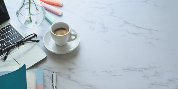 陶瓷咖啡杯放在大理石纹理的桌子上，周围是电脑笔记本电脑，眼镜，笔记本，笔记，钢笔，记号笔和植物在玻璃花瓶。图片下载