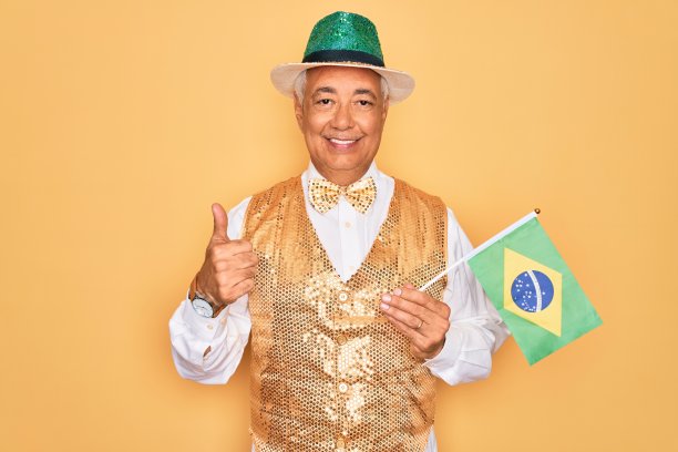 白发苍苍的中年男子穿着巴西狂欢节的习俗，手捧巴西国旗开心地笑着做ok手势，用手指竖起大拇指，做优等手势图片下载